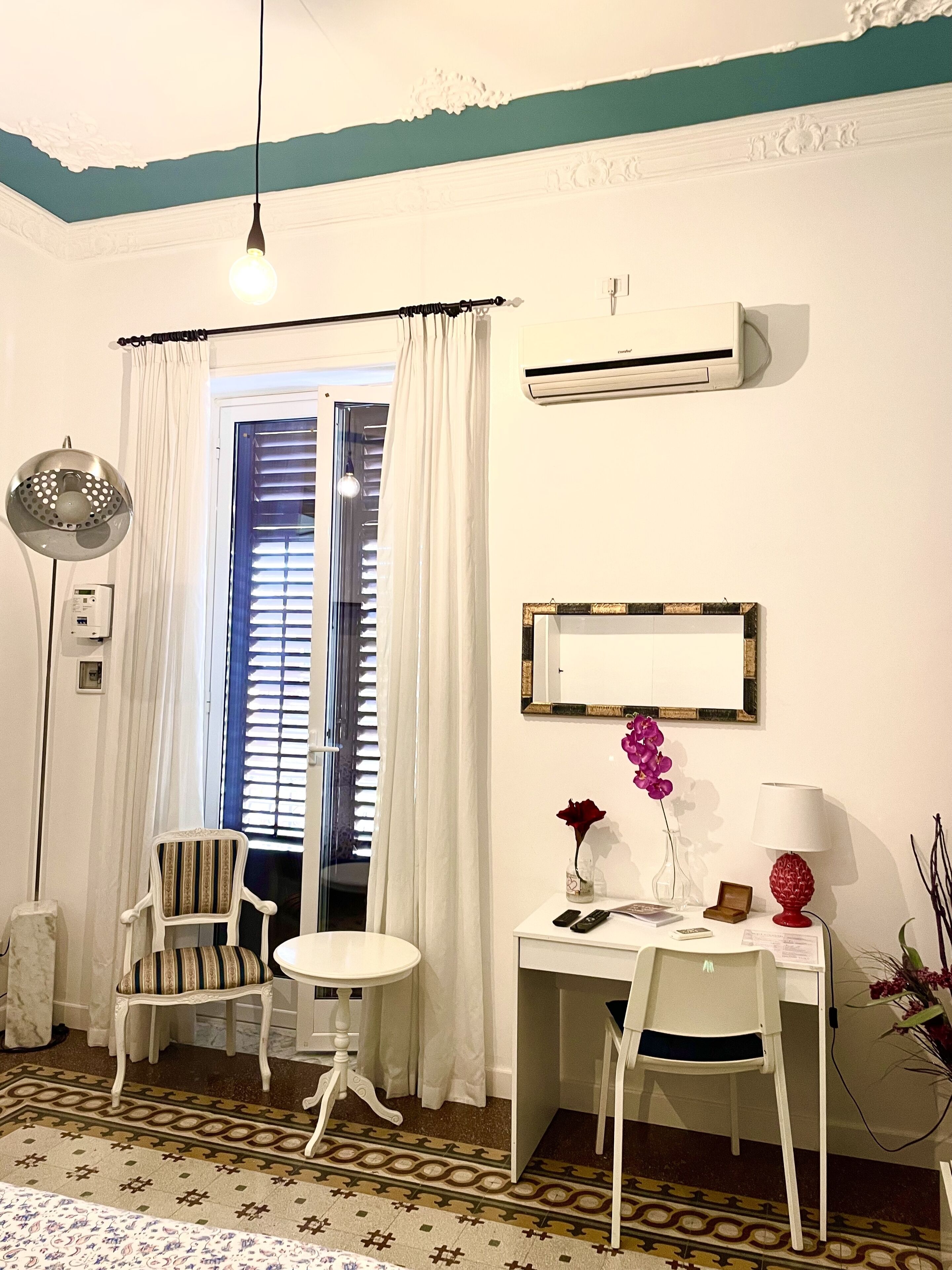 A room with a white desk, two chairs, a small table, a lamp, a mirror, and a window with curtains.