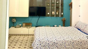 A bedroom with a teal accent wall, a large bed with a floral bedspread, a flat-screen TV mounted above the bed, and a small bedside table with a lamp and decorative items. - Funky flat (Palermo)