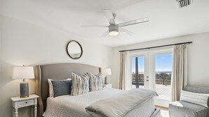 Een slaapkamer met een bed, bedlampen, een plafondventilator, een raam met gordijnen en uitzicht op de omgeving.