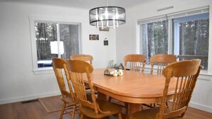 A dining room with a wooden table and six chairs, large windows, and a chandelier. - Sebec Lake House (Dover-Foxcroft)