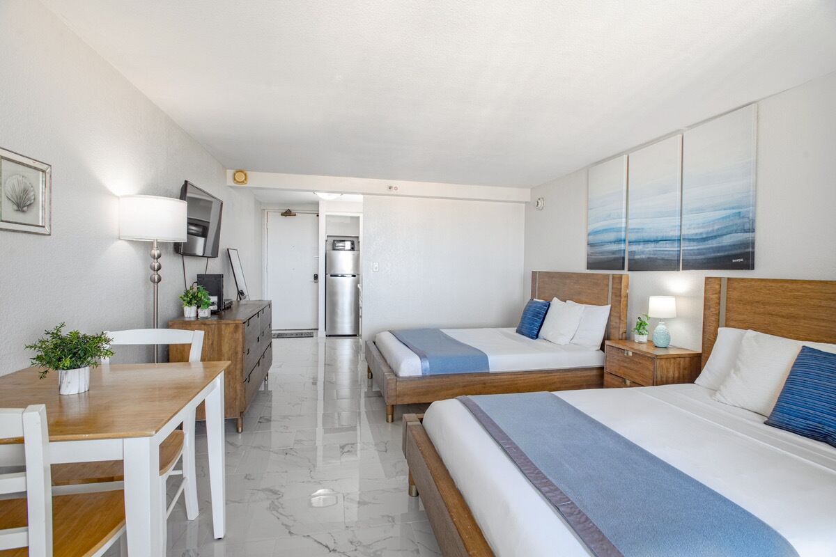 A hotel room with two beds, a desk, a chair, a refrigerator, and a TV.