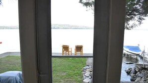 Une vue à travers une fenêtre sur un lac, avec deux chaises en bois sur une terrasse, un objet couvert sur la gauche et un bateau amarré près des rochers.