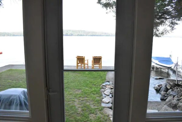 Une vue à travers une fenêtre sur un lac, avec deux chaises en bois sur une terrasse, un objet couvert sur la gauche et un bateau amarré près des rochers.