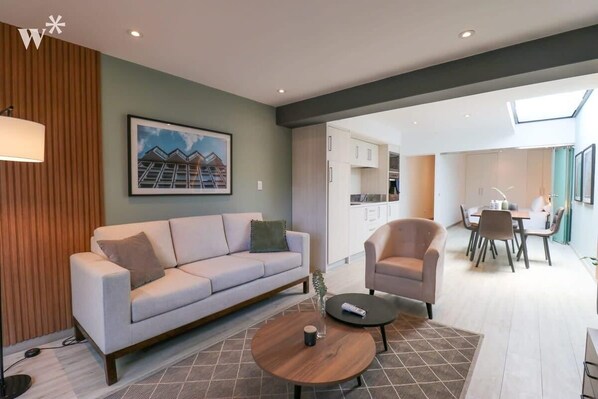Una sala moderna con sofá, sillón y mesa de centro. Se ve un comedor con mesa y sillas al fondo.