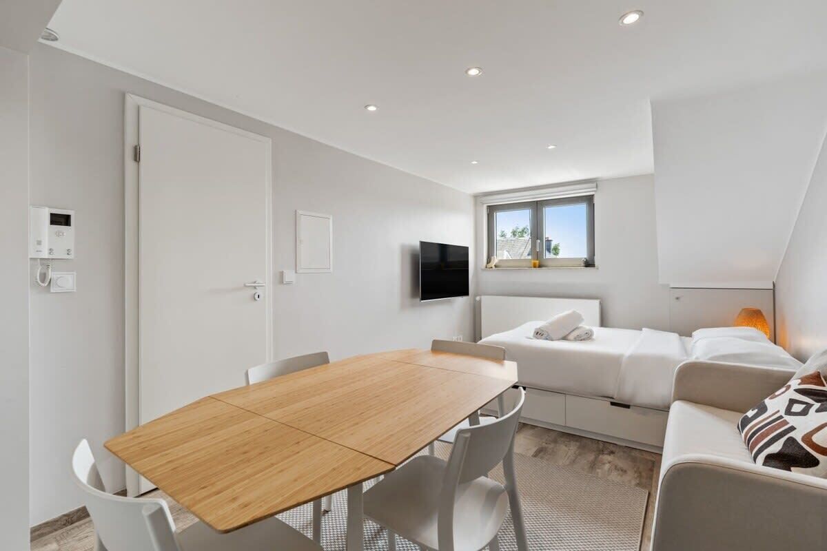 A modern, well-lit room with a wooden dining table, white chairs, a white sofa, and a flat-screen TV mounted on the wall.