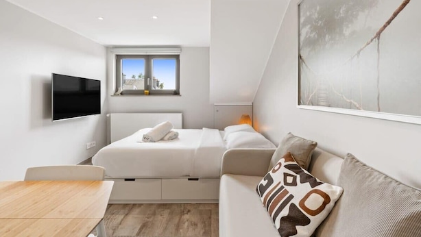 Una habitación moderna y bien iluminada con una cama, un sofá, un televisor y una ventana con vistas a edificios.