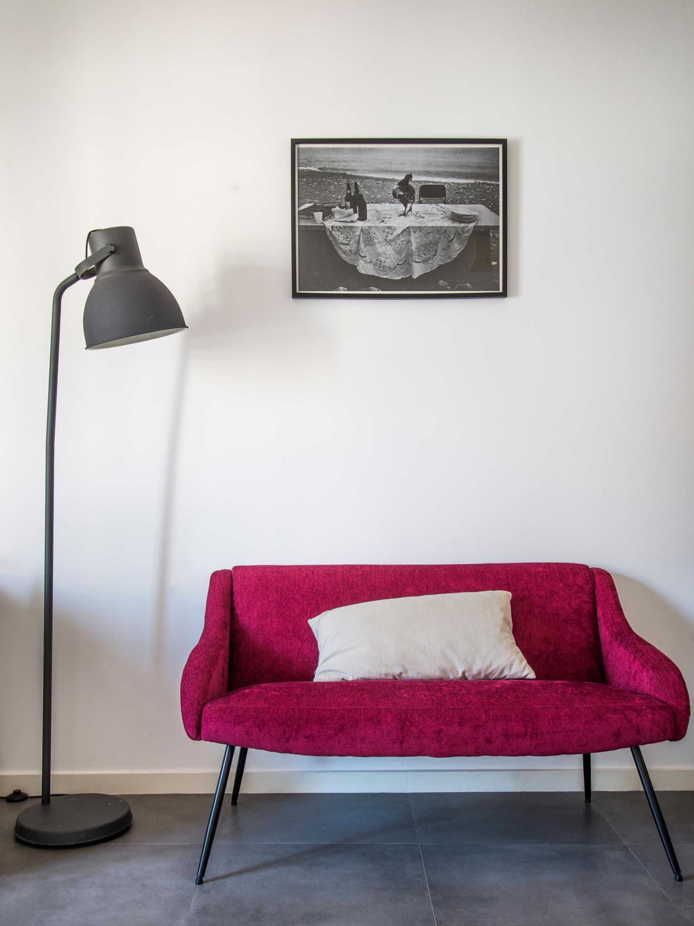 A red sofa with a white pillow, a black floor lamp, and a black and white photograph on the wall.