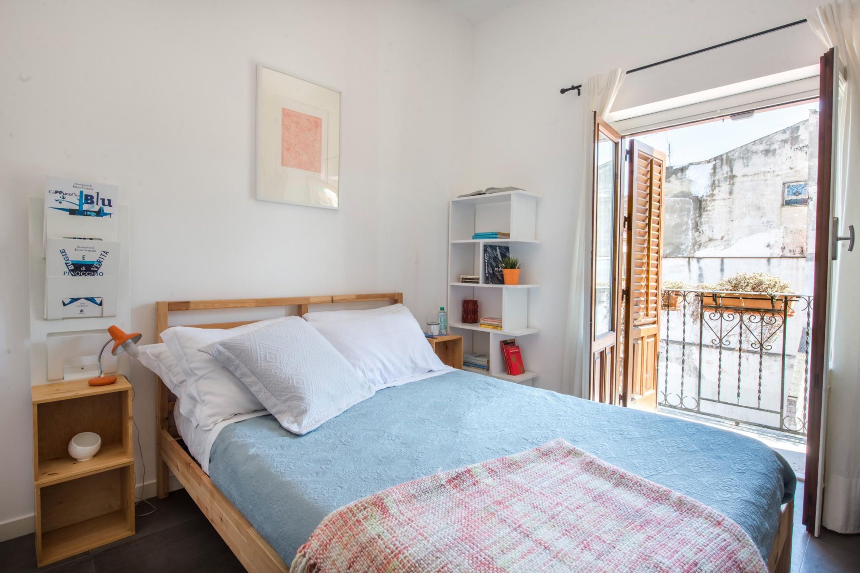 A bedroom with a bed, a bedside table, a shelf with books and a plant, and a view of the outside balcony.