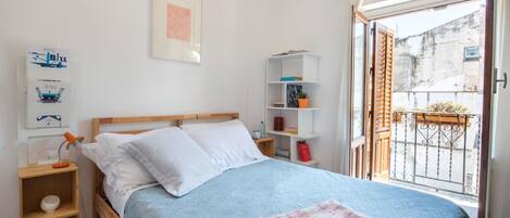 A bedroom with a bed, a bedside table, a shelf with books and a plant, and a view of the outside balcony.