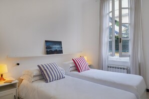 A hotel room with two beds, a nightstand with a lamp, a painting on the wall, and a window with curtains. - A Pochi Passi da Piazza de Ferrari (Genoa)