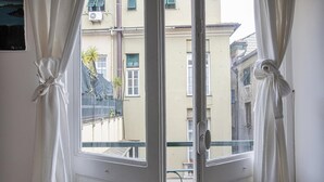 A view through a window with white curtains tied back, revealing a balcony with a metal railing and a building facade in the background. - A Pochi Passi da Piazza de Ferrari (Genoa)