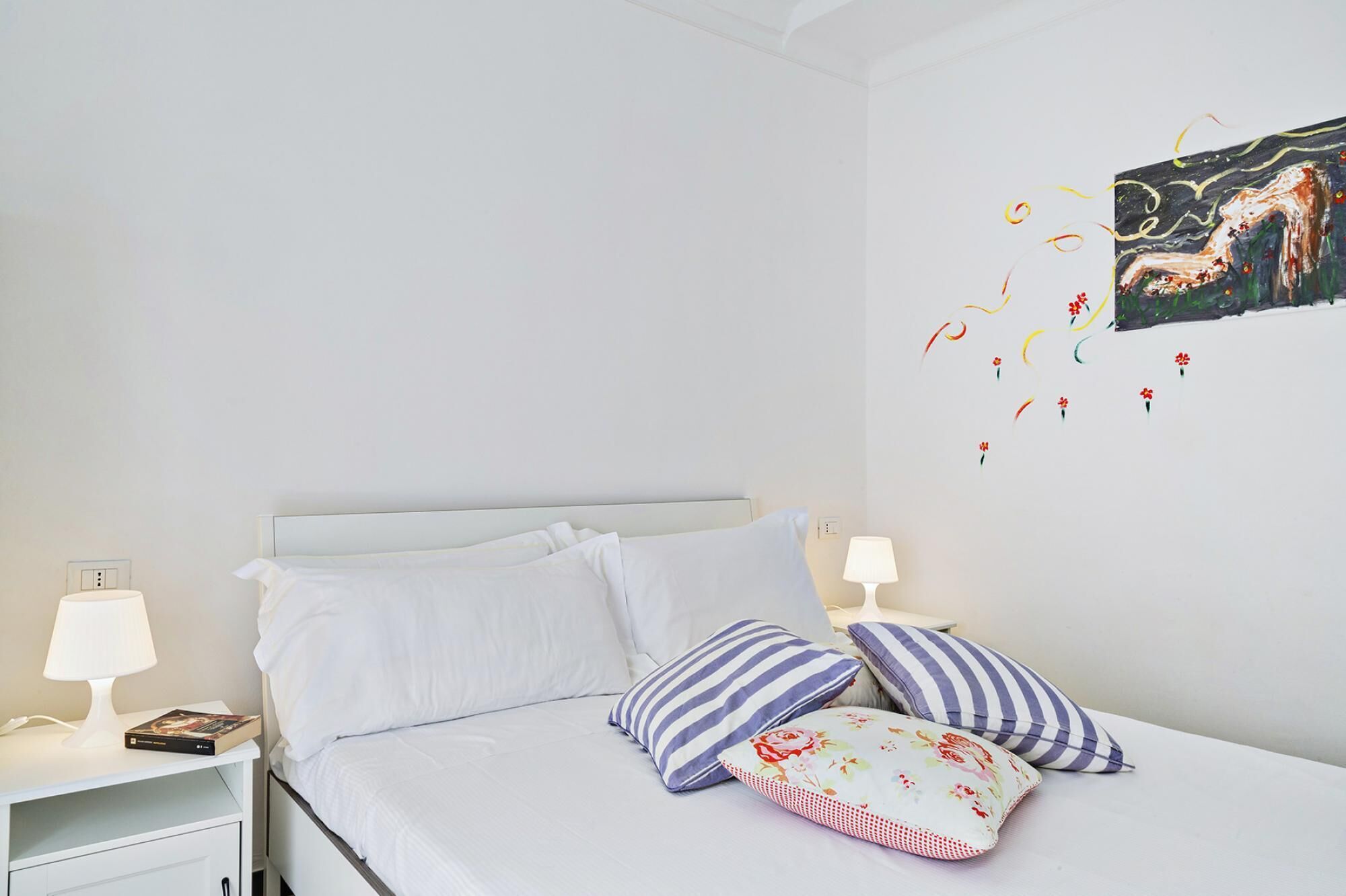 Un lit bien fait, avec une literie blanche et des oreillers à rayures, une table de chevet avec une lampe et un élément décoratif mural.
