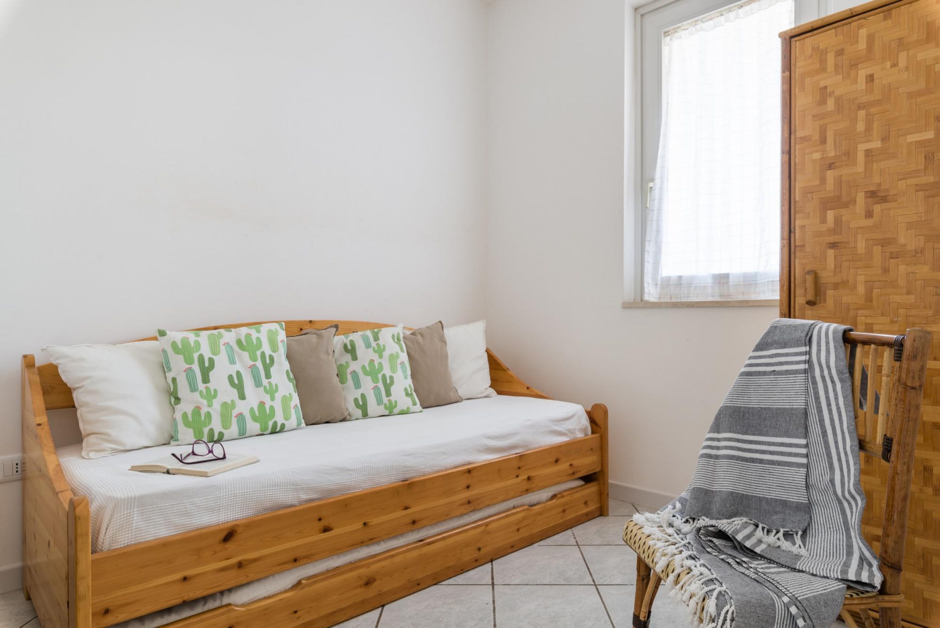 Un letto in legno singolo con materasso bianco e cuscini a motivi di cactus, una piccola sedia di legno con coperta a righe e un armadio in legno con motivo a spina di pesce.