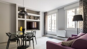 A modern living room with a purple sofa, a dining table with chairs, a TV on the wall, and a large window with patterned curtains.
