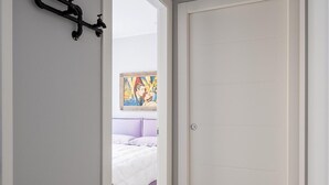 A hallway with a white door, a bed with purple bedding, and a painting on the wall.