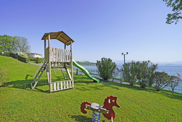 A playground with a wooden slide and climbing structure, set against a backdrop of a lake and clear sky.