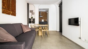 A modern living room with a gray sofa, a dining area with a round table and chairs, a kitchen with a stone backsplash, and a mounted TV. - San Michele Apartments - Lapis by Wonderful Italy (Bologna)