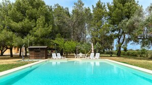 Una piscina rectangular con agua azul cristalina, rodeada de césped y árboles. Se ven dos sillas reclinables blancas y una pequeña estructura de madera al fondo.