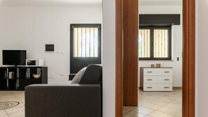 Una sala moderna con un sofá gris, un televisor, un mueble blanco pequeño y una ventana con persianas.