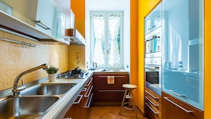 Une cuisine compacte équipée d’une cuisinière à gaz, de deux éviers, d’une fenêtre avec des rideaux et d’un petit tabouret.