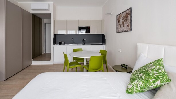 A modern hotel room with a bed, green chairs, a small table, and a kitchenette in the background. - Studio Green al Parco by Wonderful Italy (Bologna)