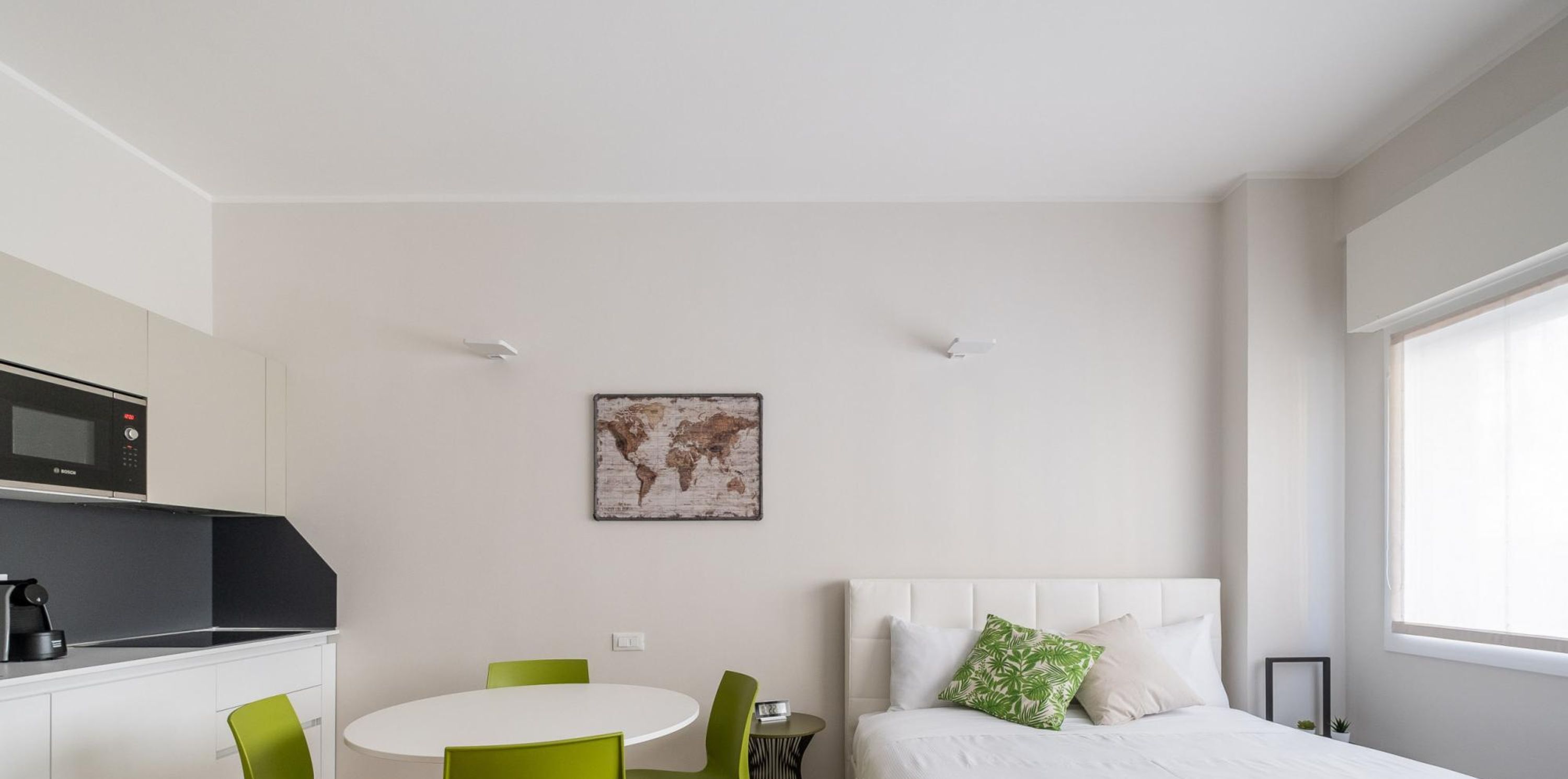 A modern hotel room with a kitchenette, a round dining table with green chairs, a bed with white bedding, and a world map artwork on the wall.