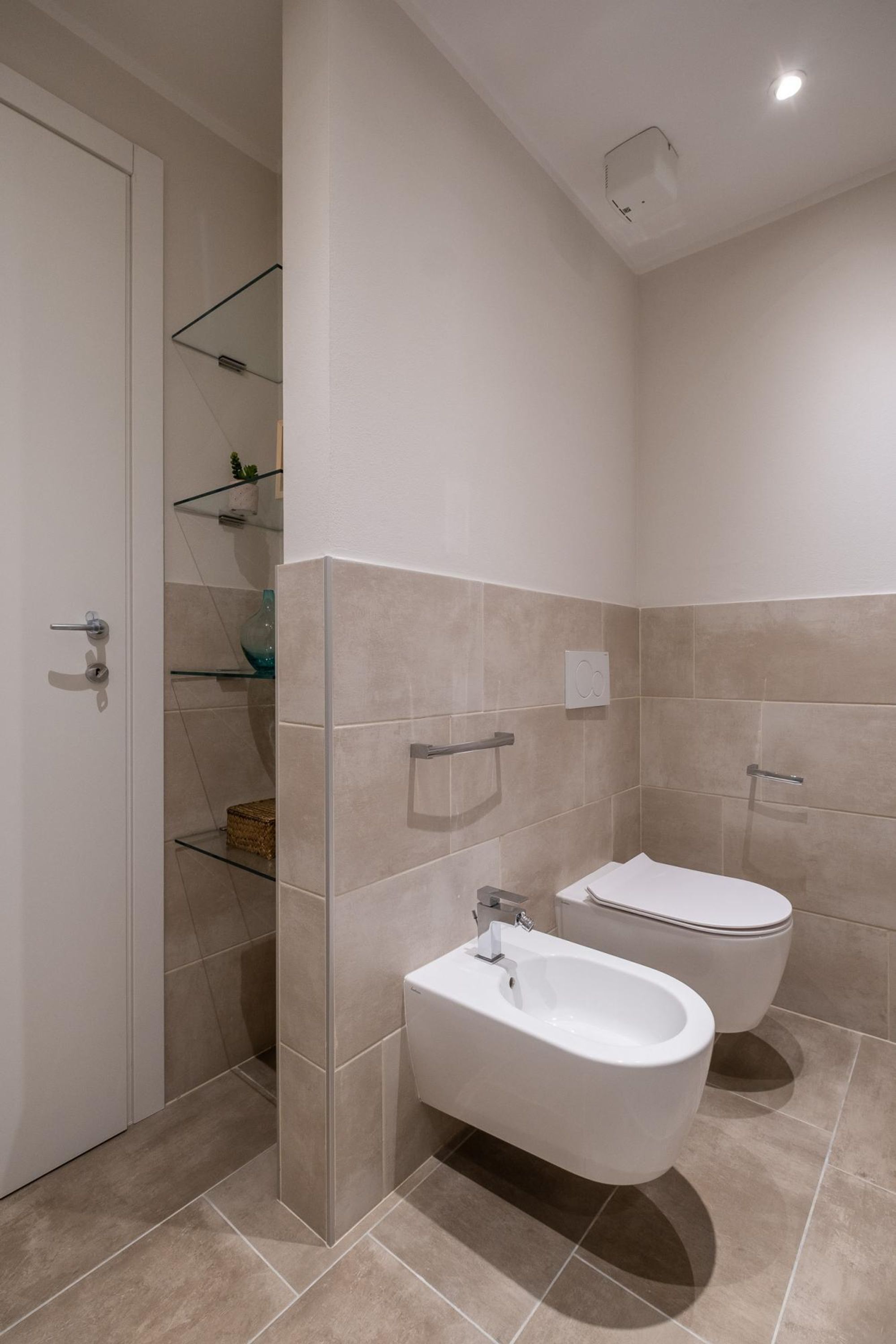 Une salle de bain moderne équipée d’un bidet, de toilettes et d’une étagère en verre où sont disposés des objets décoratifs.