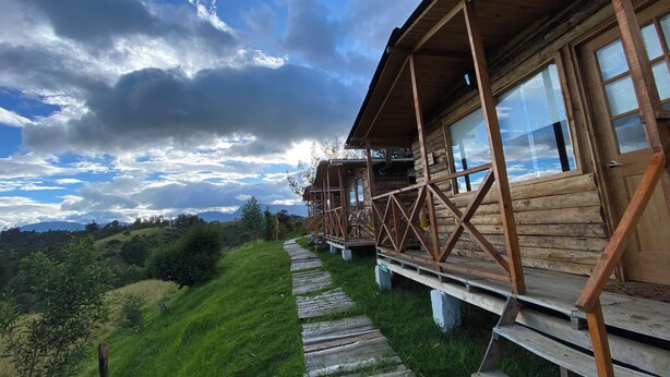 Une cabane en bois dotée d’une terrasse en bois qui mène à un porche, le tout entouré d’une végétation luxuriante et avec une chaîne de montagnes en arrière-plan.