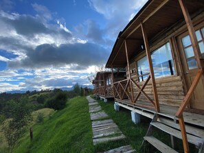 A wooden cabin with a wooden deck leading to a porch, surrounded by lush greenery and a mountainous backdrop. - Cabañas el refugio del santo (Guasca)