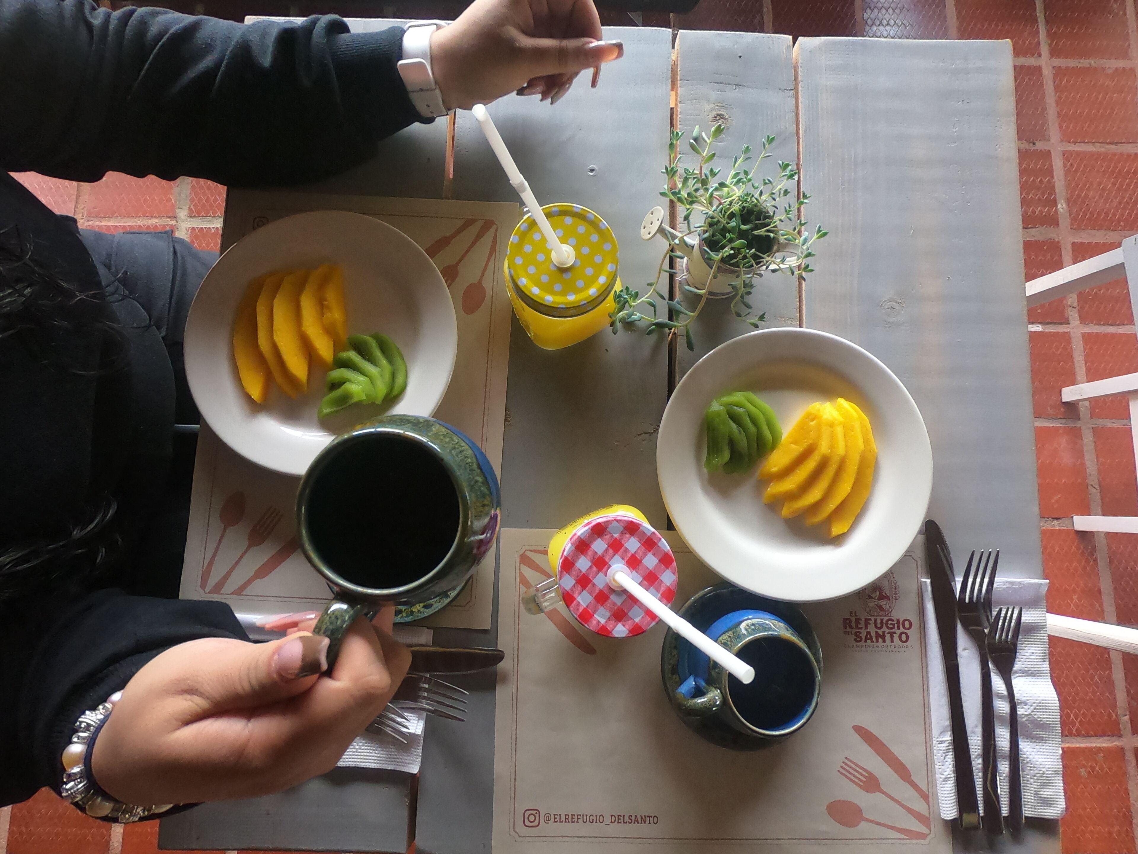 A table with plates of sliced mango and green mango, a yellow polka-dotted cup, a small potted plant, and a menu.