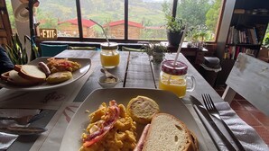 Une table de petit-déjeuner avec des assiettes contenant des œufs brouillés, du bacon, des toasts et un hot-dog. On y trouve deux verres de jus d’orange et un pot de confiture. La table est placée dans une pièce d’où l’on peut admirer un paysage rural à travers de grandes fenêtres.
