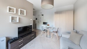 A modern living room with a flat-screen TV, a dark wooden cabinet, a dining area with a table and chairs, and a large wardrobe. - Residenza Miralago With Pool - Ground Floor Studio Apartment (Desenzano del Garda)
