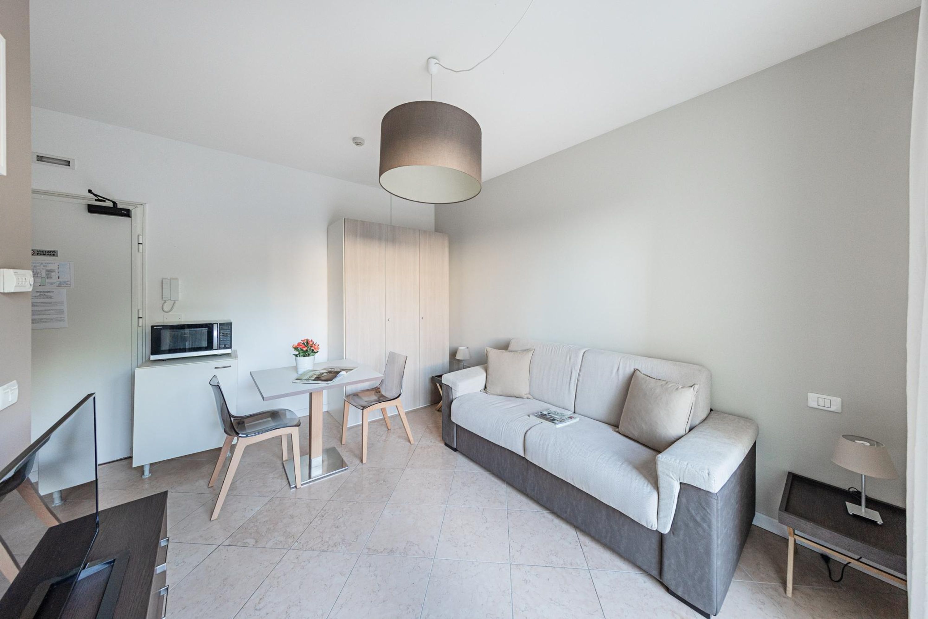 A modern, well-lit living room with a grey sofa, a dining table with chairs, a kitchen area with a microwave, and a hanging light fixture.
