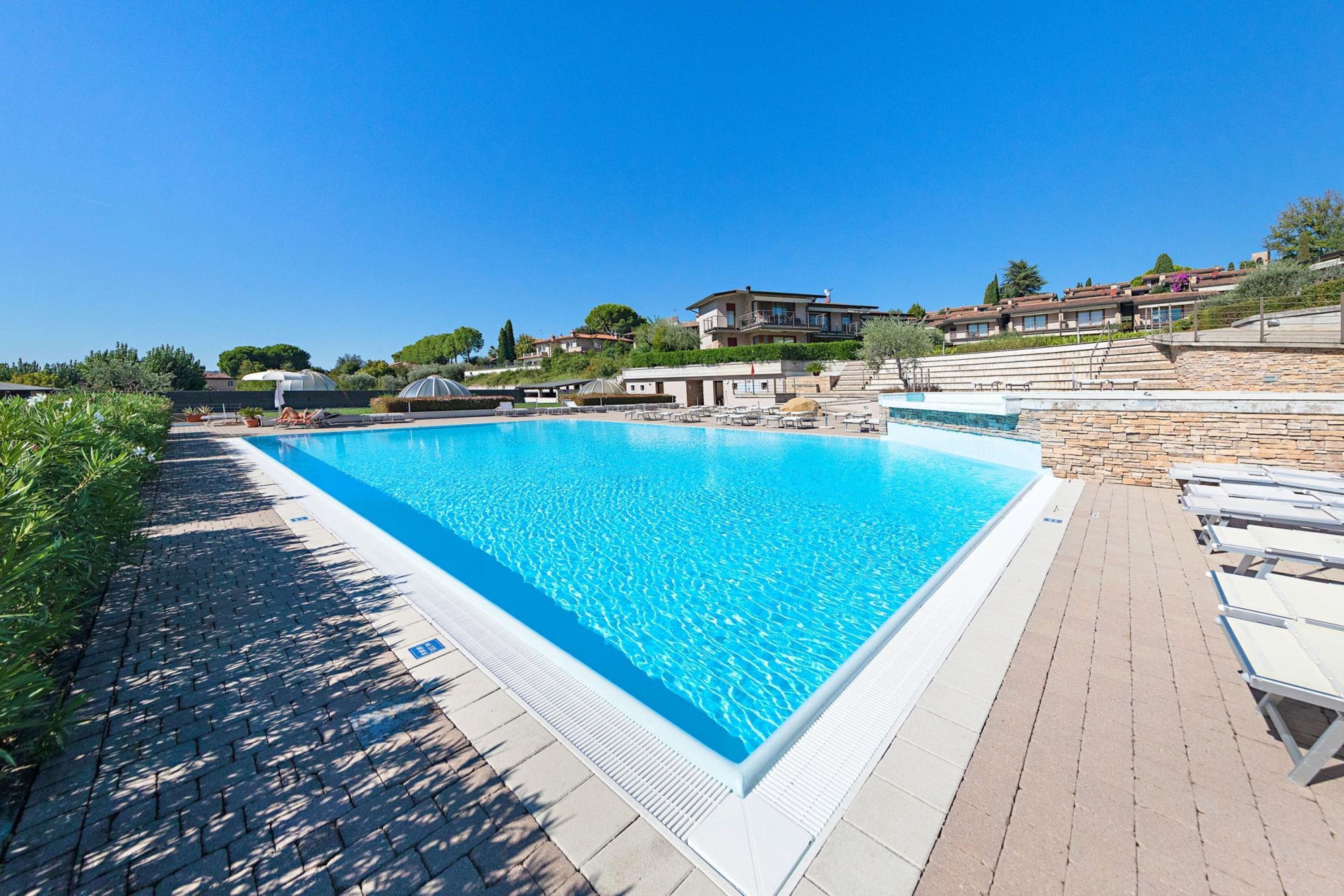 Ein rechteckiger Swimmingpool mit gefliestem Sonnendeck, umgeben von Liegestühlen und einer Steinmauer.