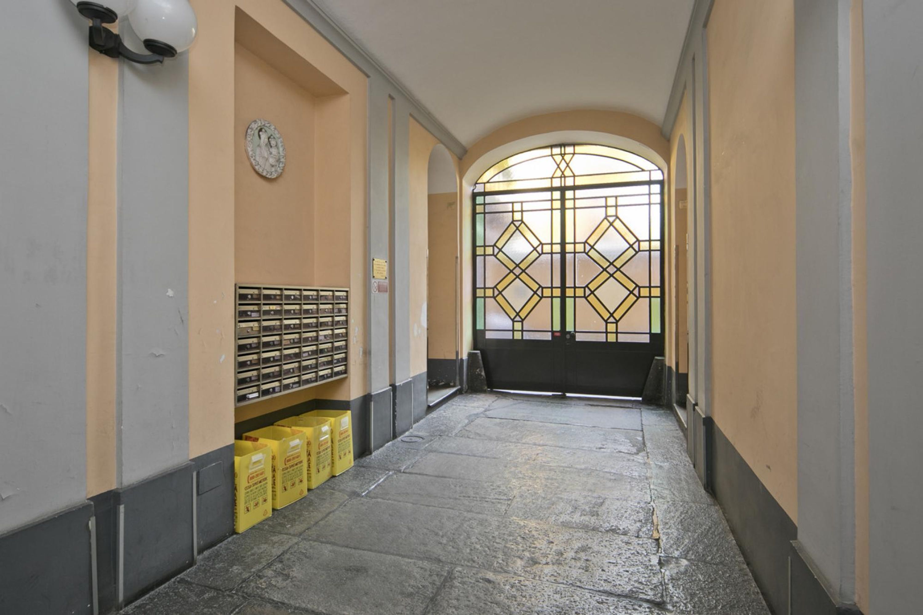 Ein Korridor mit Fliesenboden, einer Uhr an der Wand und einer Tür mit geometrischem Buntglasdesign.