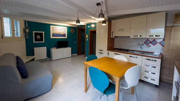 A modern kitchen with white cabinets, a wooden dining table, and blue chairs. There is a sofa and a television in the living area.