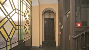 A narrow hallway with a dark wooden door, a glass door with geometric patterns, and a metal gate on the right.