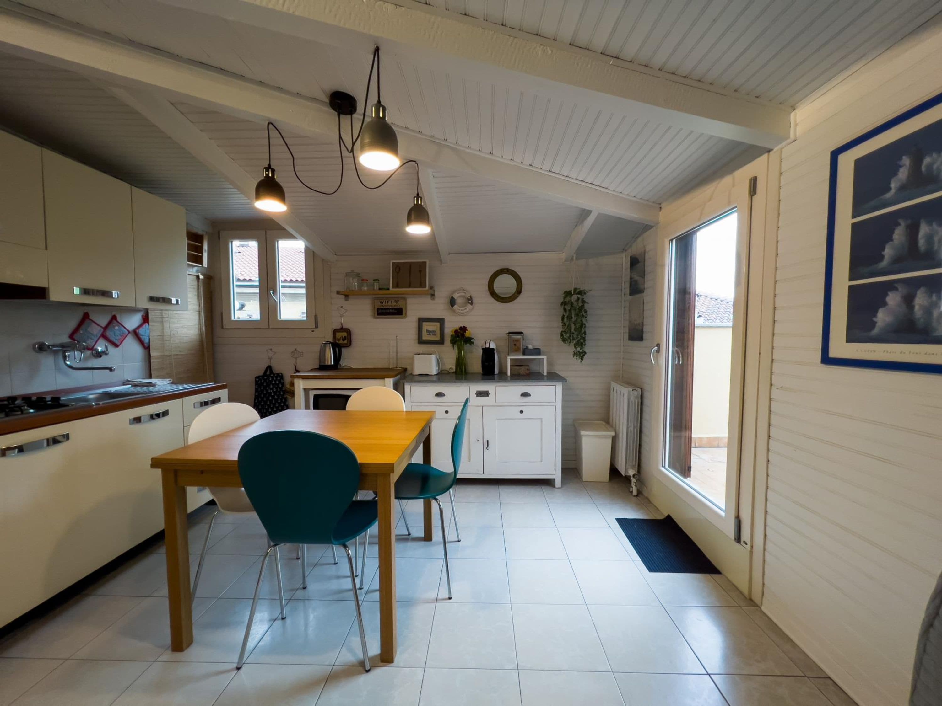 Una cucina compatta con zona pranzo, caratterizzata da un tavolo in legno, sedie color petrolio e lampade a sospensione.