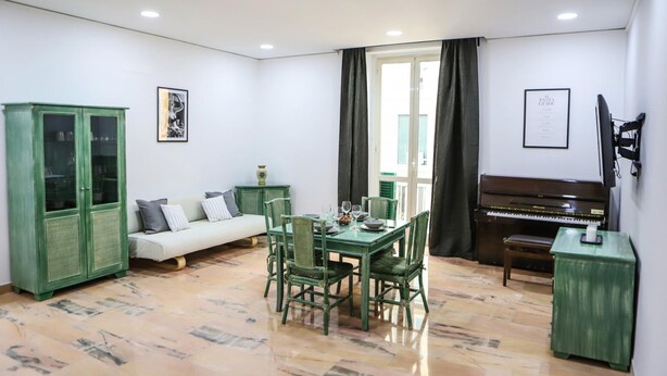 Een woonkamer met een groene eettafel, groene stoelen, een bank, een groen kastje, een piano en een raam met gordijnen.