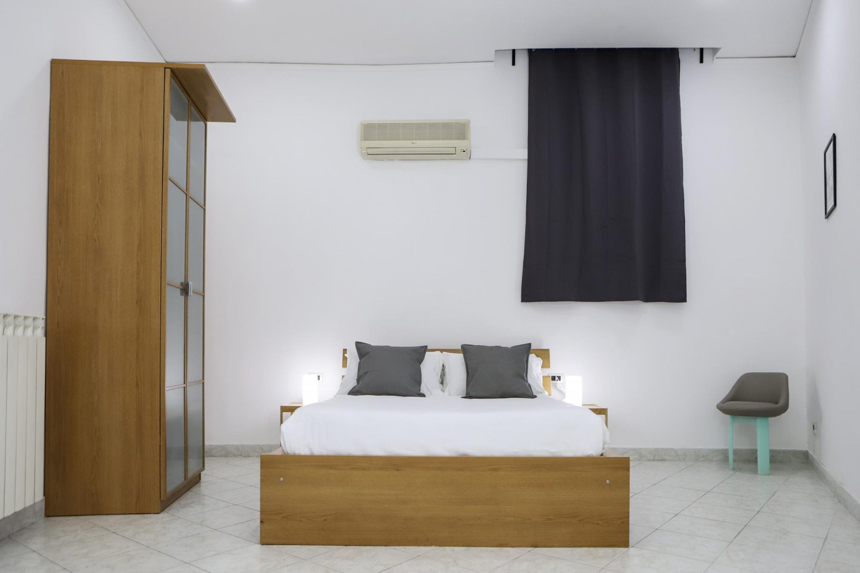 Une chambre avec un lit en bois, une table de chevet, un petit tabouret, un climatiseur fixé au mur et une porte coulissante.