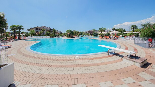 A large outdoor swimming pool with a curved design, surrounded by lounge chairs and umbrellas, and a building in the background. - Mandorli 1 18 Apartment by Wonderful Italy (Desenzano del Garda)