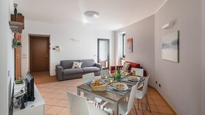 Un salon moderne avec un canapé gris, une table à manger dressée pour deux personnes et un coin cuisine visible au fond.