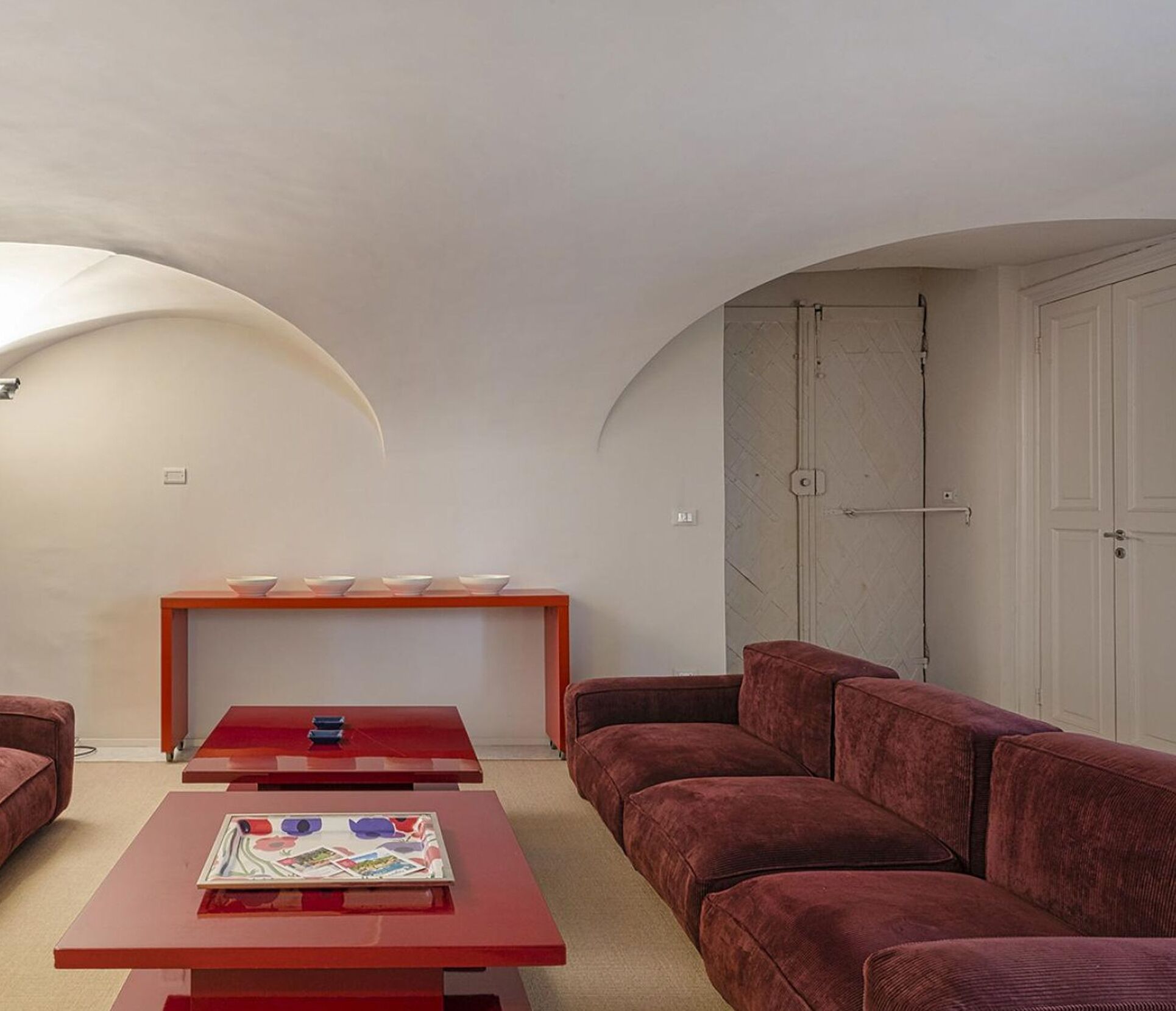 Ein Wohnzimmer mit einem roten Sofa, einem roten Couchtisch und einem roten Beistelltisch. Im Hintergrund eine Holzbänkl mit Schalen darauf und eine weiße Tür.