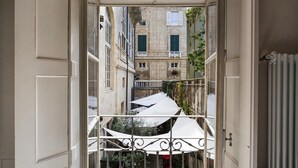 Un balcon ouvert avec des auvents blancs, une vue sur une rue étroite bordée d’immeubles et un radiateur fixé au mur.