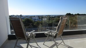 Two wooden lounge chairs with cushions on a balcony with a glass railing, overlooking a coastal town.
