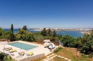 Área de piscina com espreguiçadeiras e guarda-sóis, cercada por vegetação e com vista para o horizonte costeiro.