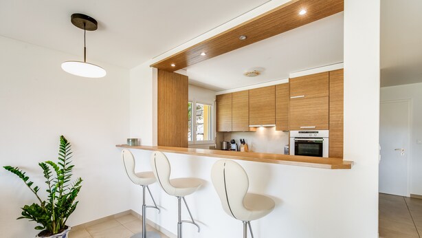 A modern kitchen with wooden cabinets, a built-in oven, and a bar area with three bar stools.
