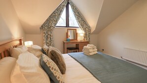 A cozy bedroom with a sloped ceiling, a bed with pillows, a bedside table with a lamp, a stack of towels, and a window with curtains. - Beechside (Ambleside)