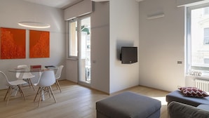 Een moderne woonkamer met een eethoek, een flatscreen televisie aan de muur en een groot raam dat zorgt voor daglicht.