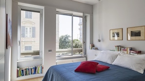 Een slaapkamer met een bed, een rode kussen, een boekenkast en twee ramen met uitzicht op gebouwen.